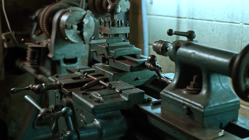 A close view of a large dark metal lathe machine in a workshop shows its various gears handles and mechanical components illuminated by a light source to the right