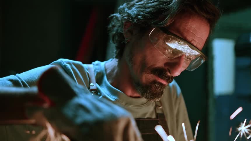 A male artisan with a beard and glasses wearing a light colored shirt and a dark apron intently works on a project that creates bright orange sparks with dark tools and a dark background in the workshop