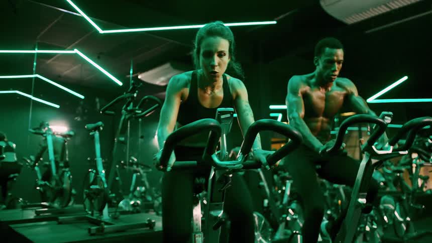 A couple both sweating and showing intense effort ride stationary bikes in a gym with green neon lights