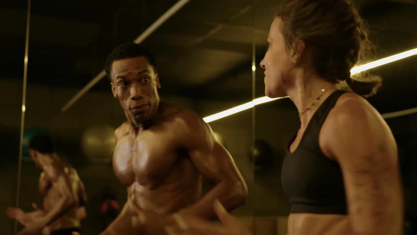 Two athletic individuals train together in a gym exchanging focused expressions during an intense workout session under warm lighting