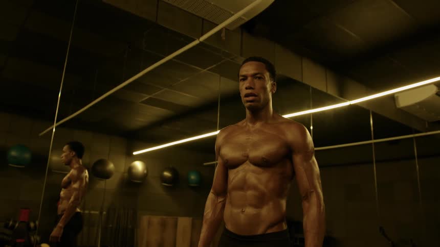 A muscular black man standing shirtless in a gym his skin glistening with sweat under warm lighting with a serious and focused expression