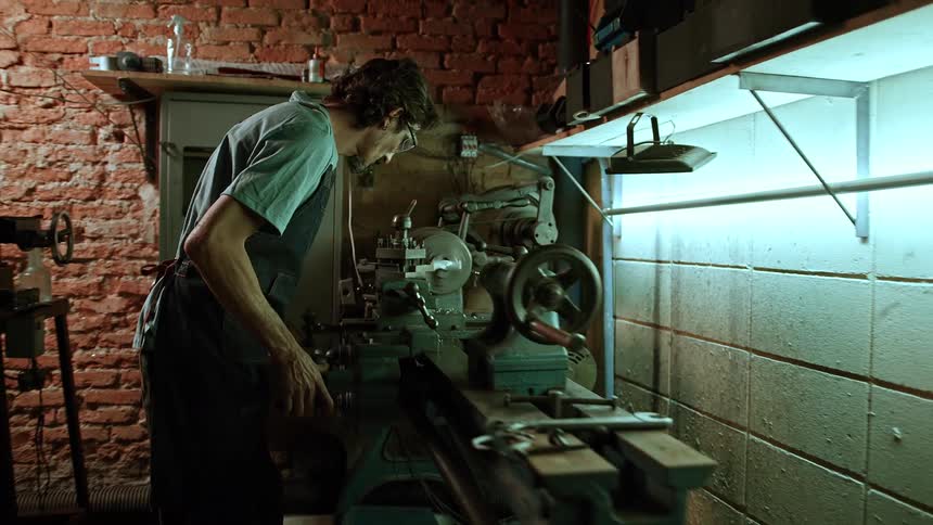 A man in a workshop is intently focused on operating a vintage metal lathe under bright fluorescent lighting with a brick wall in the background