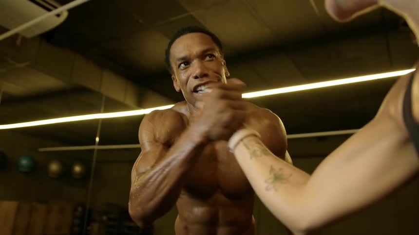 Two athletic individuals in a gym midhandshake or friendly competition with the muscular man smiling and shirtless and the woman showing butterfly tattoos on her arm