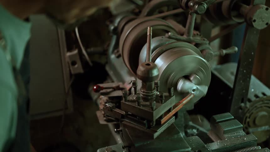 An adult male artisan with a beard and glasses wearing a light colored shirt leans over a large metal lathe machine intently focusing on the metal piece being worked