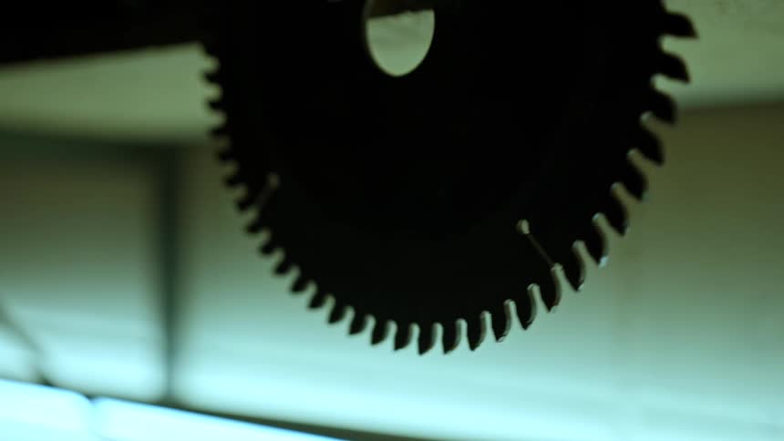 A close view of a large dark circular saw blade with sharp teeth hangs balancing with a lighter blurred background