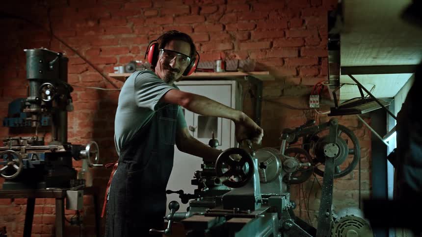 An older adult male artisan with glasses and orange ear protection wearing a light colored shirt and a dark apron operates a large metal machine in a workshop with a brick wall smiling at the viewer