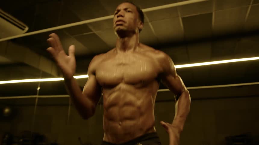 A young adult man with a muscular physique covered in sweat intensely performs a cardio exercise that involves high knees and arm movements in a gym with dark walls and horizontal strip lights on the ceiling