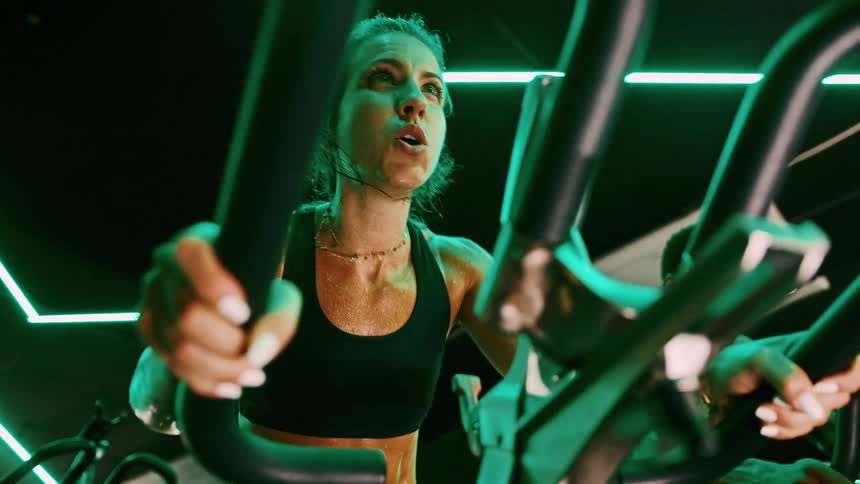 A young adult woman in a black sports bra with sweat visible on her skin works out intensely on an exercise machine looking upwards in a dimly lit room with green neon lights