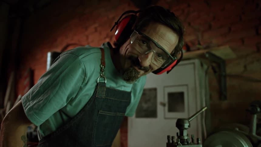 A man wearing safety goggles and earmuffs smiles while working in a workshop