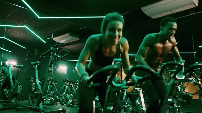 This video features a dynamic shot of a diverse group of people engaged in an intense indoor cycling class with a focus on a determinedlooking woman and a shirtless man in the foreground the dimly lit gym is accentuated by vibrant green neon lights