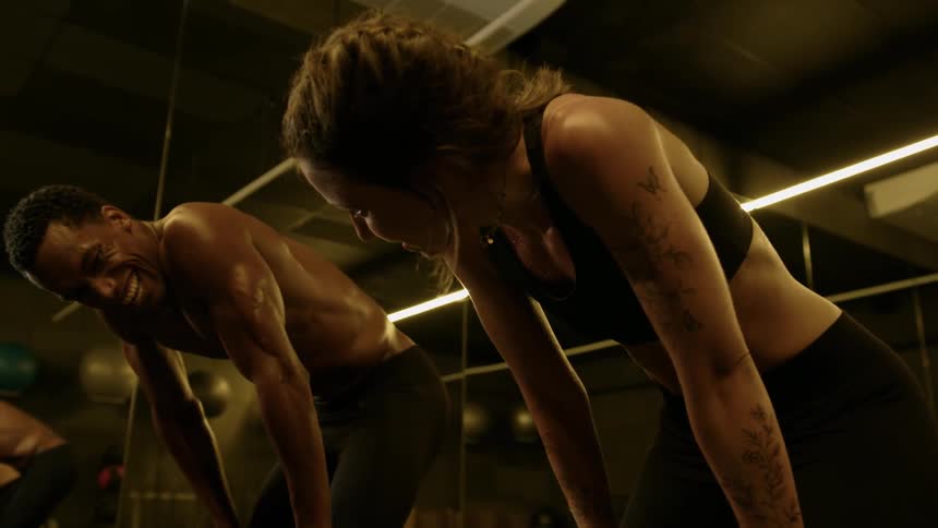 Two individuals a woman in a sports bra and a man are leaning forward with their reflections visible in a large mirror in a gym setting