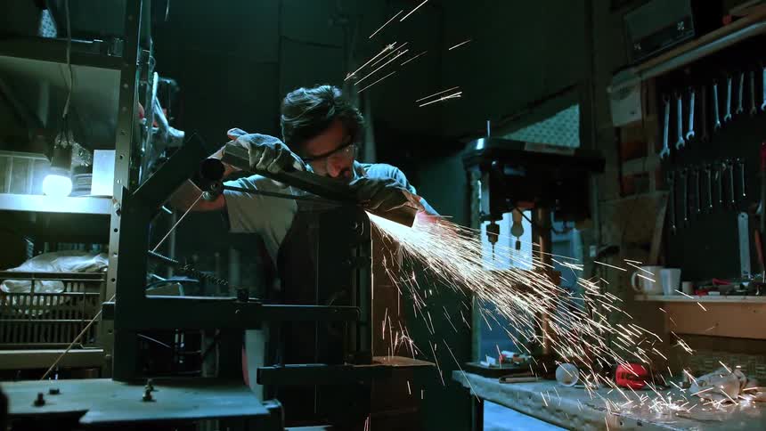 A man in safety gear grinds metal sending bright sparks flying across a cluttered workspace
