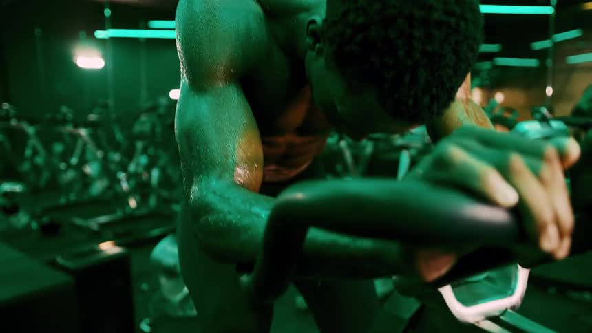A muscular black man leans forward drenched in sweat during an intense workout under green lighting