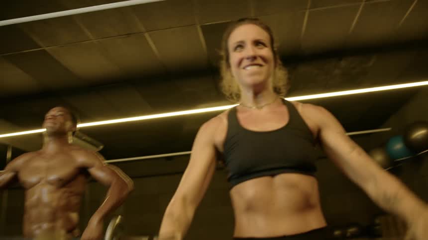 A young adult woman and a young adult man both sweating and showing intense effort lift barbells over their heads in a gym