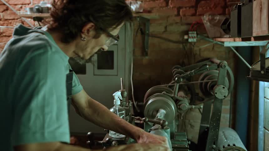 An older adult male artisan with glasses and long hair wears an apron over a light colored shirt operating machinery in a workshop with brick walls and various tools