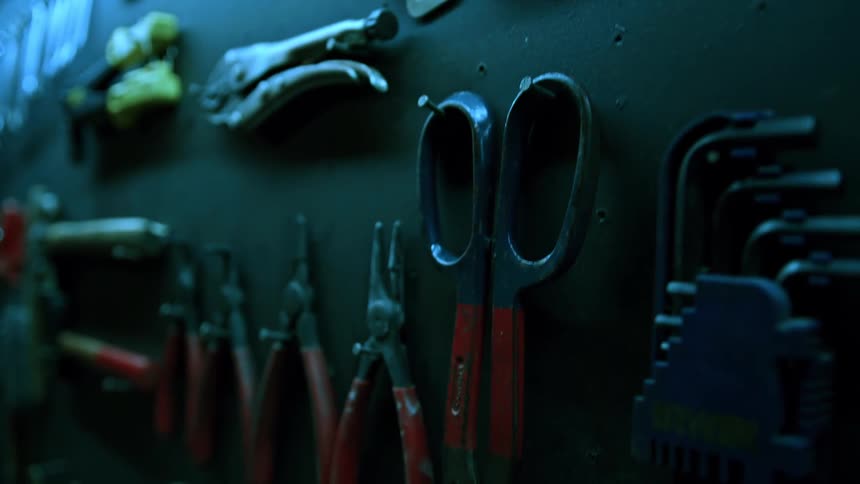 A close view of a dark tool board shows various metal tools including wrenches pliers with red handles and scissors hanging neatly arranged under dim lighting