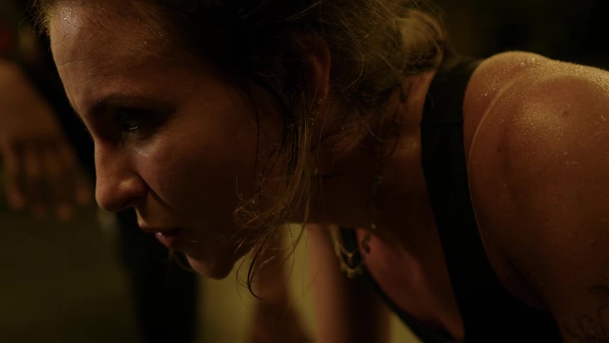 A close view shows a woman with sweat covering her face and neck looking down with an expression of intense effort wearing a dark top and dangling earrings