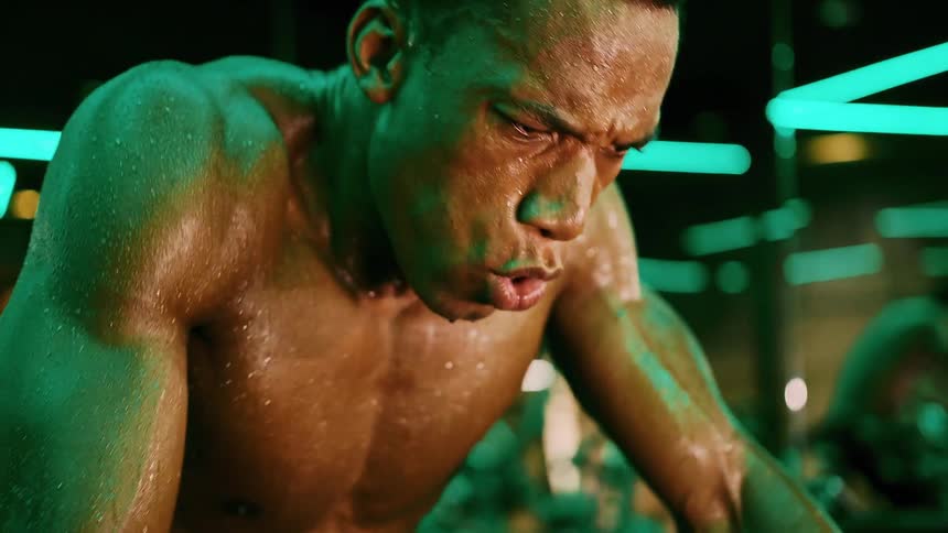 A man with a muscular physique covered in sweat looks up with an expression of intense effort and a grimace on his face illuminated by green and blue neon lights in a gym