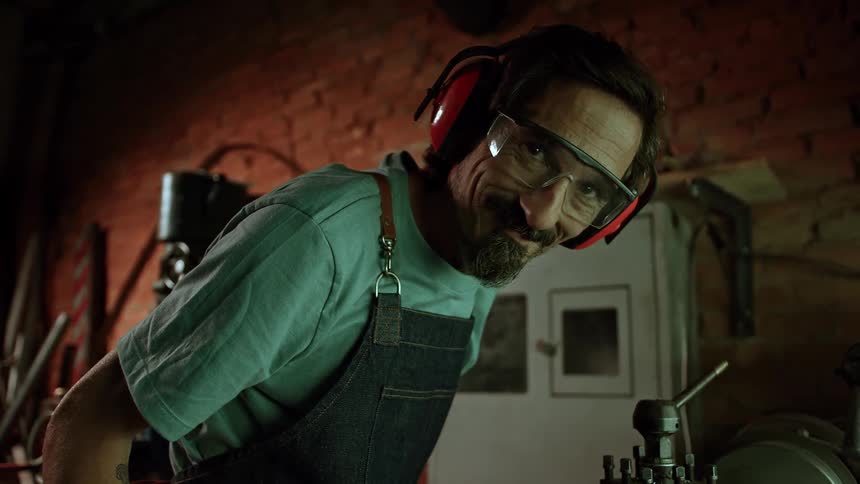 A man inside a workshop wears headphones and safety glasses while working with tools