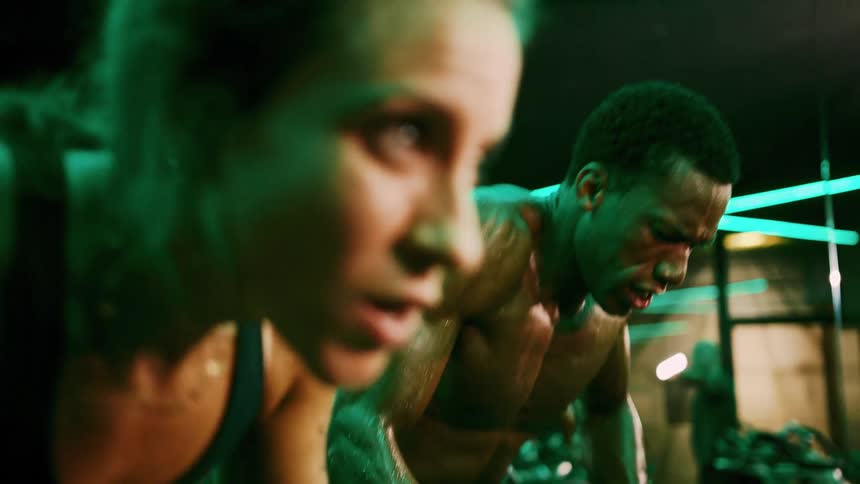 A woman in a dark sports bra and a man with no shirt on both covered in sweat ride stationary bikes with intense expressions in a gym with green neon lighting