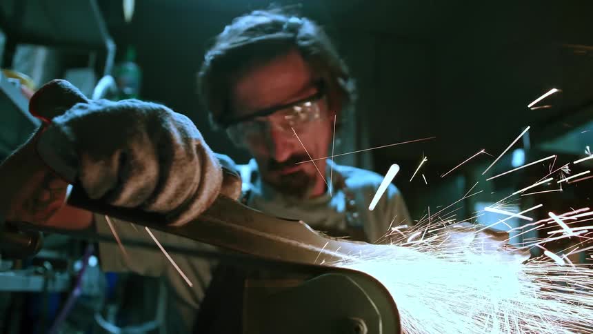 An artisan wearing safety glasses and a work glove uses a grinding tool that produces bright sparks intently focusing on the metal object in his hands