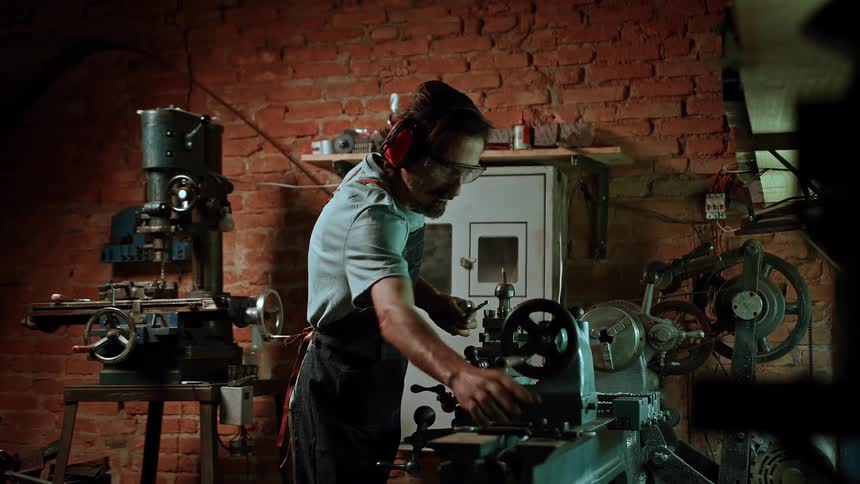 An older adult man an artisan wearing glasses and orange ear protection works on a large metal lathe in a dimly lit workshop with a brick wall