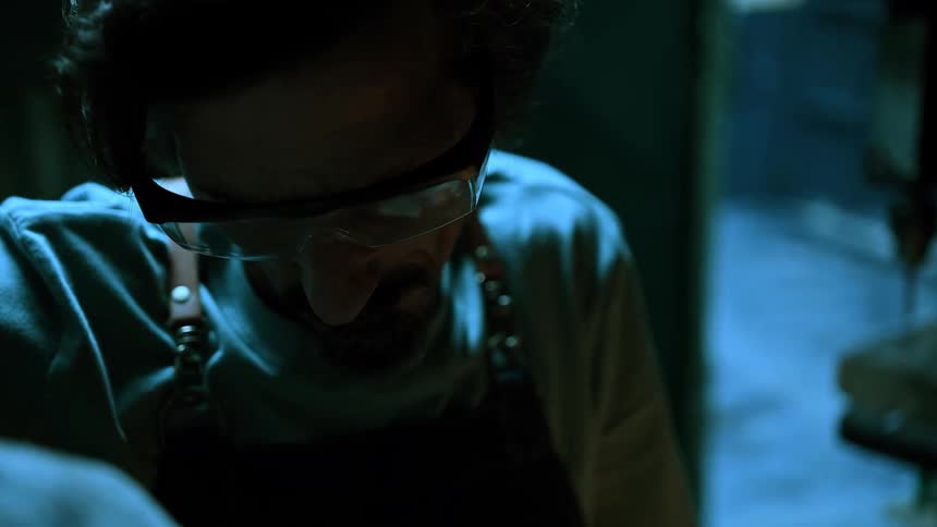 A man in a leather apron and safety glasses focuses intently on a task with sparks in a dimly lit workshop with blue ambient light