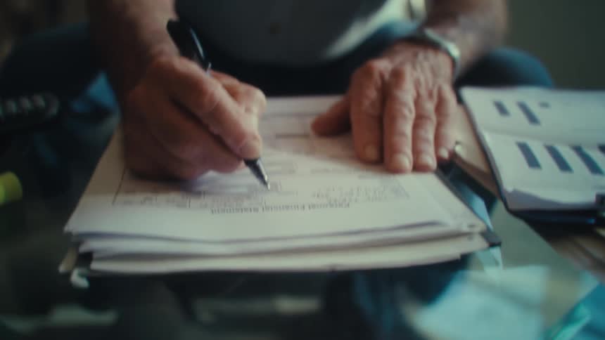 An elderly man wearing glasses and a light colored shirt sits at a table and uses a pen to write on a stack of financial documents