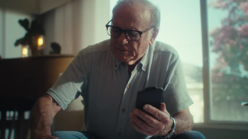 An elderly man wearing glasses sits near a sunlit window focused intently as he uses a smartphone and writing on a piece of paper
