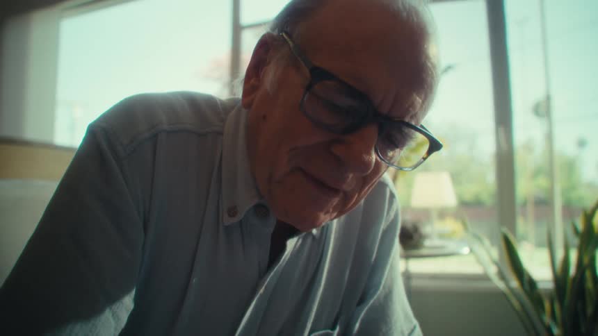 An elderly man sits in his living room at midday writing on a document when he suddenly begins to laugh