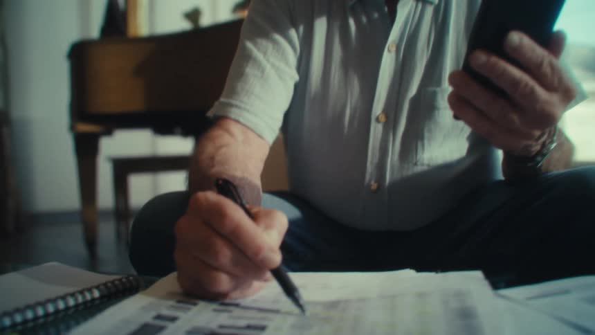 An elderly man sits with his phone in his hand while marking a paper nearby