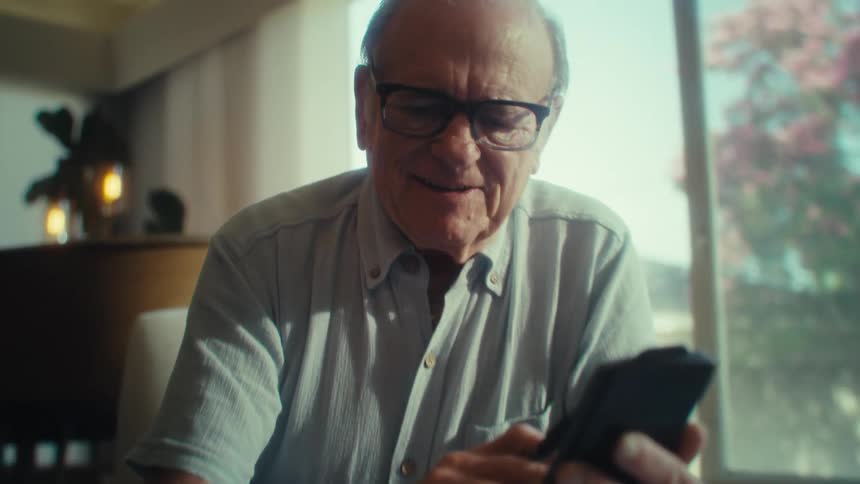 N elderly man smiles while using a smartphone in a sunlit living room
