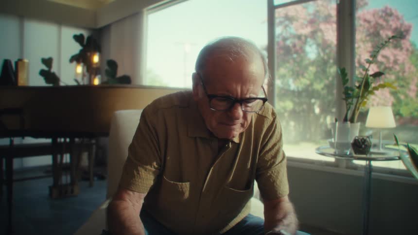 An elderly man in glasses and a brown shirt leans forward in a sunlit living room focused on an unseen task in front of him