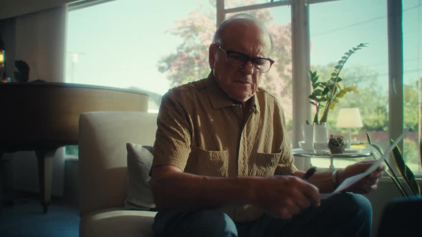 An elderly man wearing glasses sits on a chair in his living room at midday working on a laptop and looking through documents