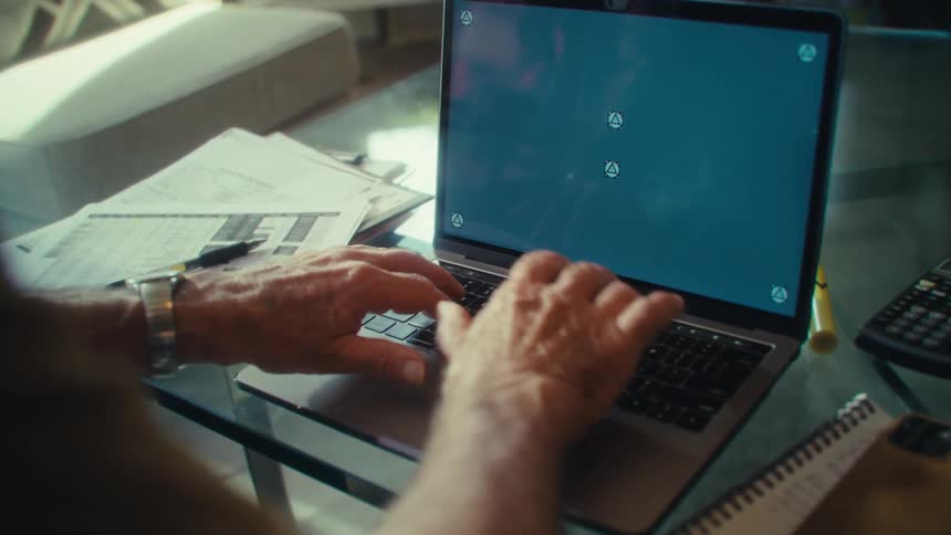 An older adult with a watch on his wrist sits at a table intently typing on a laptop with a blue screen surrounded by papers a calculator and a notebook