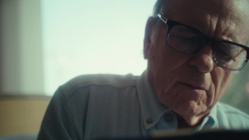 An elderly man sits in his living room at midday wearing glasses and working on a laptop