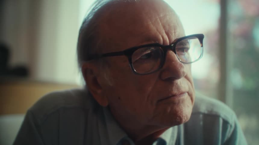 An elderly man wearing glasses sits indoors in thought then suddenly begins to smile