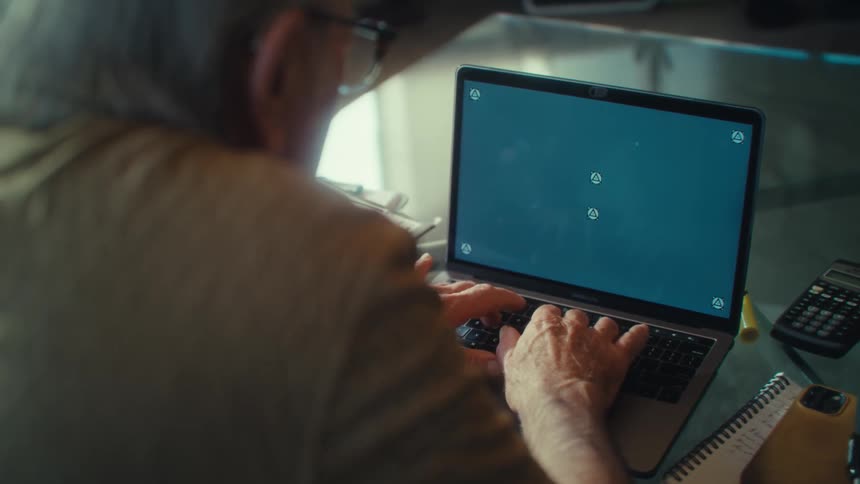 An elderly man sits indoors and types on a laptop