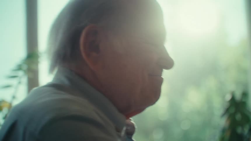 An older adult man with light colored hair and glasses is seen in profile celebrating looking down and smiling slightly with bright light coming from a window behind him