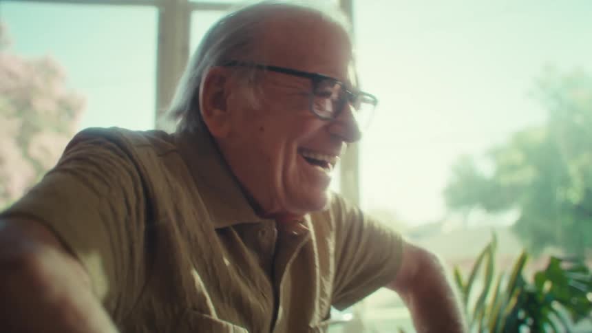 A cheerful elderly man with glasses is happily engaged with his laptop sitting comfortably on a sofa in a brightly lit room