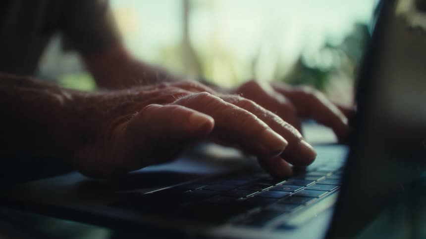 An older adult types on a laptop in a bright room filled with natural light and plants