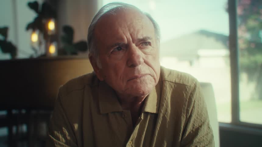 An elderly man sitting with his laptop takes off his glasses and looks up as he contemplates
