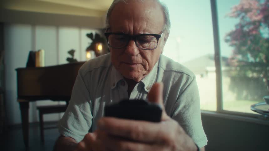 An older adult man wearing glasses and a light colored shirt holds a document and glances between it and a smartphone with a grand piano and a large window in the background of his home