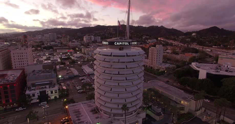 An aerial view shows the cylindrical Capitol Records building in Hollywood with an American flag flying from a flagpole surrounded by city streets with parked cars and other buildings all under a cloudy twilight sky