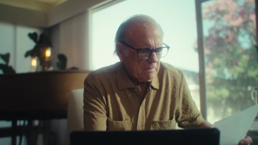 An elderly man sits in his living room wearing glasses writing on a piece of paper while looking at a laptop and examining a document