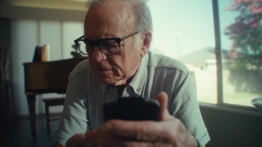 An elderly man wearing glasses sits near a window concentrating intently then laughing as he uses a smartphone in a softly lit room