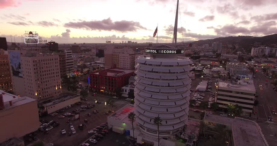 An aerial view shows the round Capitol Records building in Hollywood with an american flag waving from its rooftop flagpole surrounded by city streets and buildings under a dramatic cloudy sky