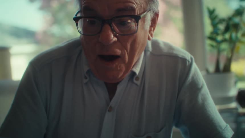 An elderly man wearing glasses looks down in surprise and smiles his mouth slightly open as he reacts to something unseen