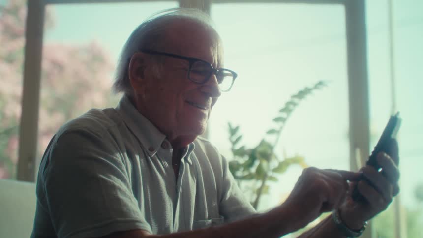 An older adult man wearing glasses and a light colored shirt sits on a couch using a smartphone interacting with the device and showing a range of emotions from concentration to a smile