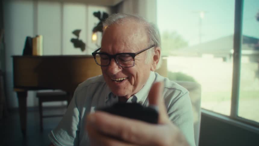 An older adult man wearing glasses and a light colored shirt holds a smartphone and reacts with a surprised then happy and laughing expression while looking at the phone in a well lit room with a grand piano and a large window in the background